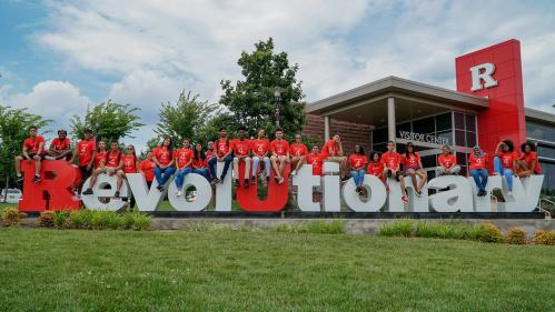 Diverse group of male and female students sitting on large red and white letters that spell out Revolutionary. 