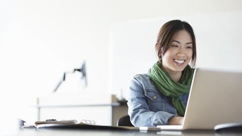 young asian woman looking at her laptop smiling. She is wearing a green scarf and denim jacket