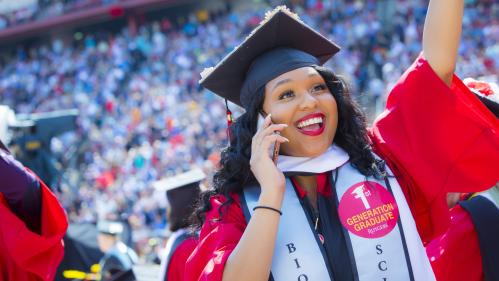 Woman in graduation cap and gown, smiling and talking on a cell phone wearing a sticker that says first generation.