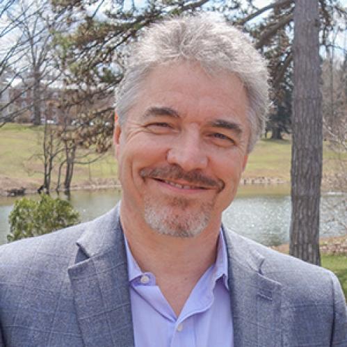 Paul Mclean has grey hair and is smiling in front of trees and a lake. He is wearing a grey suit and a blue collared shirt
