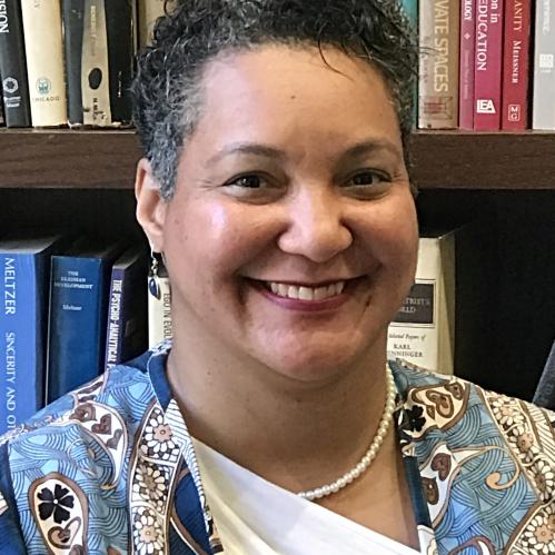 Nikol has short curly hair and fair skin. She is smiling while standing in front of a brown velvet curtain. She is wearing a leopard printed blouse and cardigan.