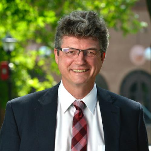 Peter March is a caucasian man with salt and pepper grey hair. He is wearing a black suit with a red plaid tie and standing in front of a green tree background