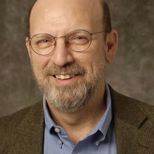 Jorge Schement is a wearing wire rimmed glasses a brown jacket and blue collared shirt. He is bald and has fair skin and a brown and grey moustach and goatie.