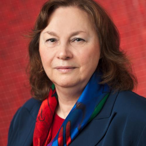 Nancy is has shoulderlength wavy brown hair, fair skin and she is smiling. She is standing in front of a Red textured wall and she is wearing a navy blue jacket, a red bouse and a multi-colored scarf