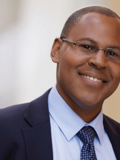 Jason Greary is an african american man with crossly cut hair. He is wearing clear rimmed glasses with a navy blue jacket and a navy blue spotted tie.