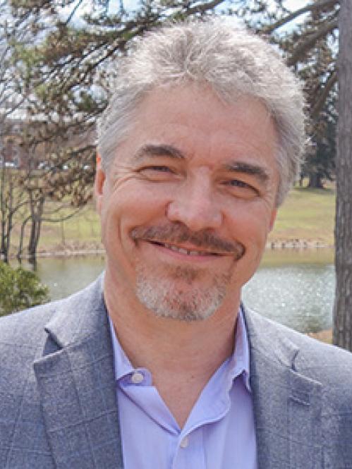 Paul Mclean has grey hair and is smiling in front of trees and a lake. He is wearing a grey suit and a blue collared shirt