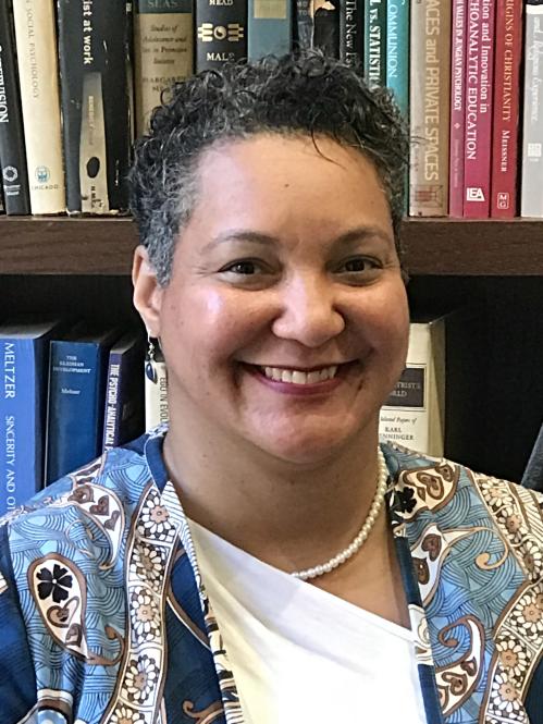 Nikol has short curly hair and fair skin. She is smiling while standing in front of a brown velvet curtain. She is wearing a leopard printed blouse and cardigan.