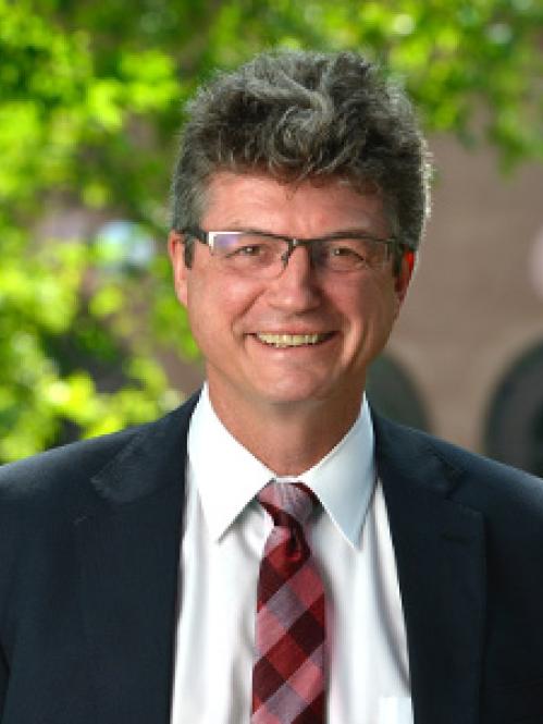 Peter March is a caucasian man with salt and pepper grey hair. He is wearing a black suit with a red plaid tie and standing in front of a green tree background