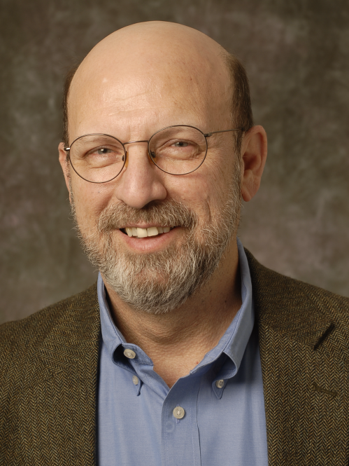Jorge Schement is a wearing wire rimmed glasses a brown jacket and blue collared shirt. He is bald and has fair skin and a brown and grey moustach and goatie.