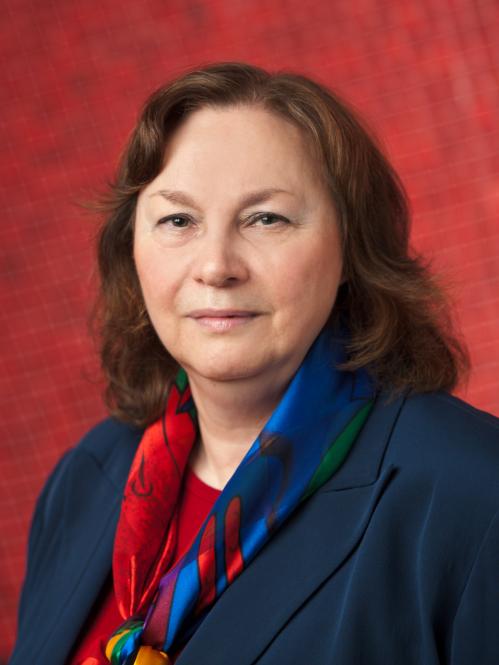 Nancy is has shoulderlength wavy brown hair, fair skin and she is smiling. She is standing in front of a Red textured wall and she is wearing a navy blue jacket, a red bouse and a multi-colored scarf