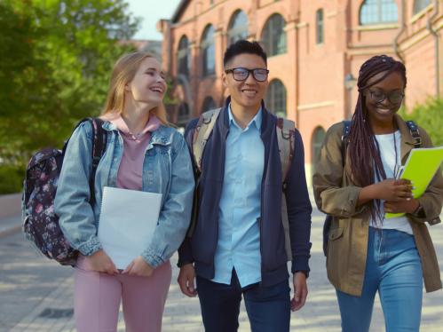3 students walking on campus talking and smiling. Left to right: young caucasian student, young asian american student, young african american student