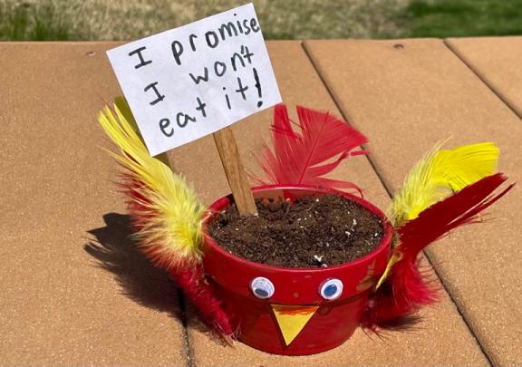A red flower pot with paper eyes, beak and feathers. With a sign that says "I promise I won't eat it!" 